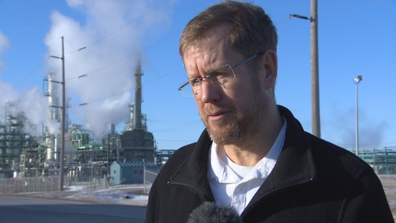 A man wearing glasses is pictured in front of a refinery.