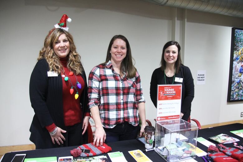 Three women dressed in festive outfits stand at a table taking donations for the food bank