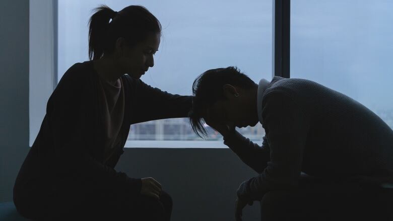 A woman is shown consoling another person, with their faces and bodies all shadowed, in front of a big window.