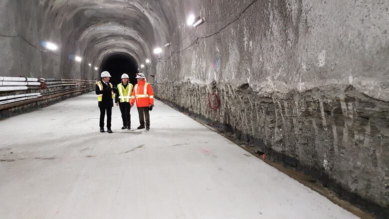 3 people stand in a brightly-lit underground tunnel. They are wearing high-viz vests and hard hats. They are about 100 feet away from the camera. The tunnel has high ceilings and goes on so you can't see the end.