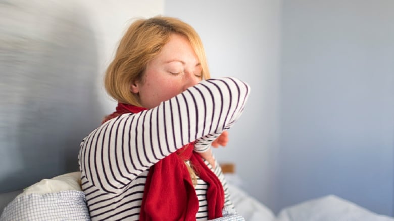 A blonde woman sits on a bed and coughs into her elbow. 