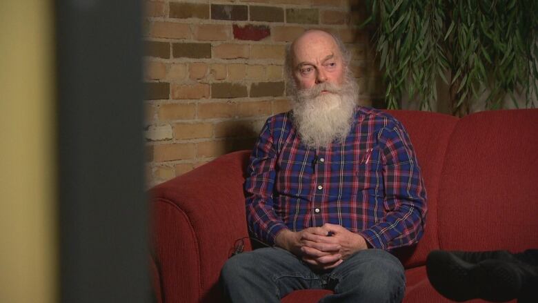 A balding man with a full white beard sits on a red couch with his hands in his lap.