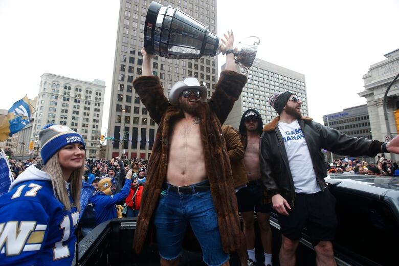 A man in fur coat and cowboy hat, with a bare chest under the open coat, holds up a trophy.