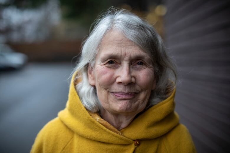 A woman with white hair and wearing a yellow hoodie smiles for the camera.