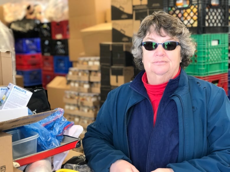 A woman with salt and pepper hair, wearing a navy blue coat, red turtle neck and dark glasses looks towards the camera. In the background are stacks of food storage crates in black, red, blue and green.