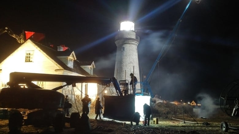 A film crew work in front of a lighthouse on a dark night.