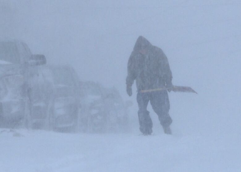 A person carrying a shovel and walking through heavy snowfall.