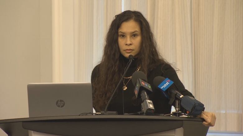 A Black woman wearing a black turtle neck stand at a podium in front of multiple mics