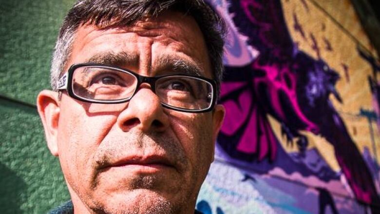 Portrait of an Indigenous man with short grey-black hair, in front of a colourful mural.