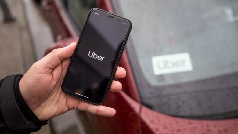 A person holds up a phone with 'Uber' on it, with a car in the background having an 'Uber' label on the dash.