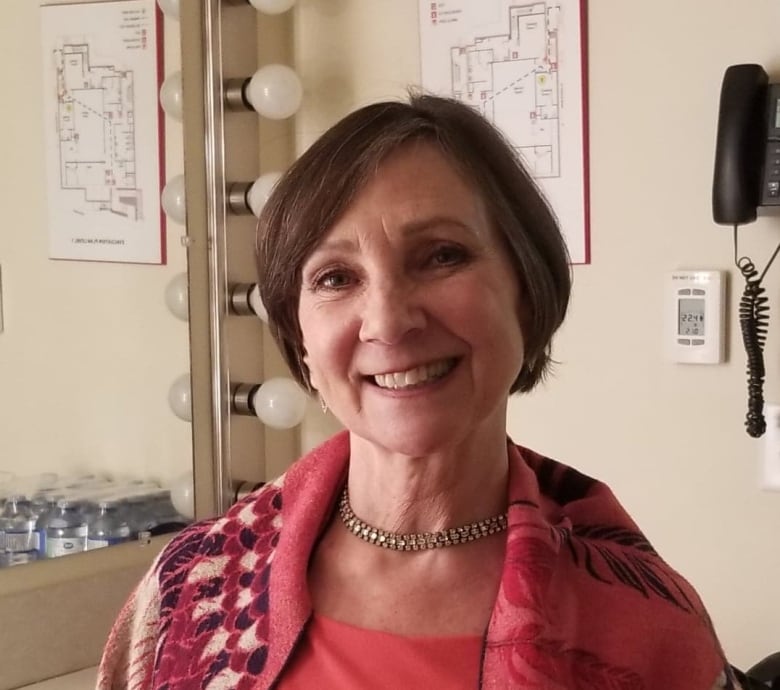 Woman with short brown hair smiling, she has a necklace and is wearing a pink sweater. She is sitting with her back against a mirror.