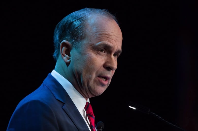 A man in a suit and red tie speaks at a microphone.