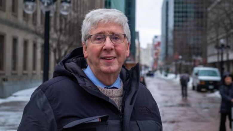 A man with grey hair and glasses stands on a snowy street and looks directly into camera. 