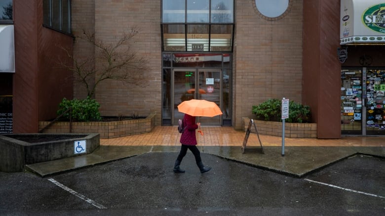 A person wearing a read coat and carrying an orange umbrella walks in the rain in front of a brick building.