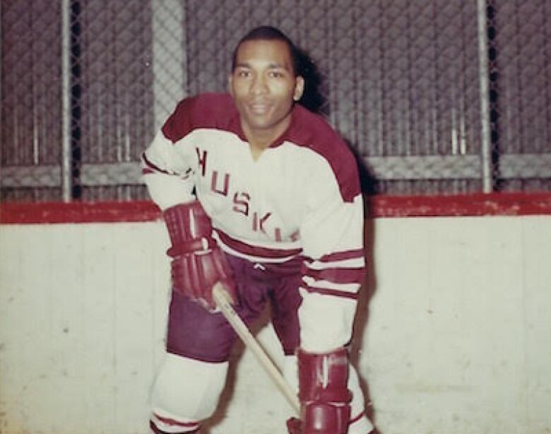 Man in a hockey rink posing for the camera. He leans forward on his stick.