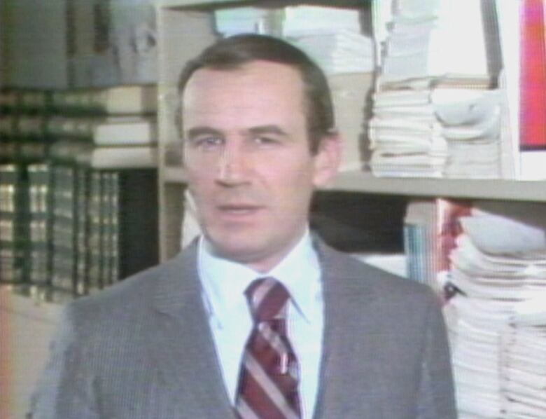 Man in jacket and tie stands in front of shelves full of books and papers