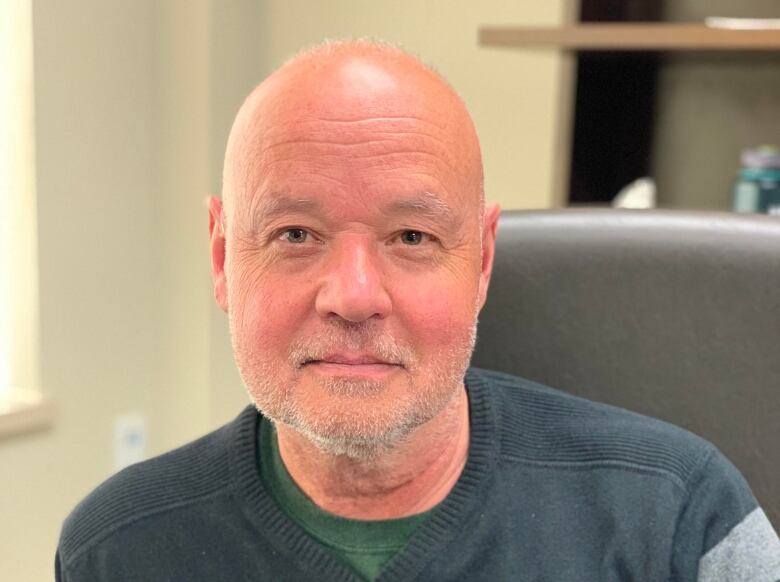 A bald man in a dark, striped sweater sits in an office chair.