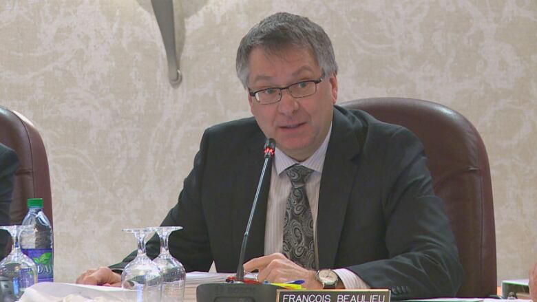 A man in a suit sits in front of a microphone in a boardroom.