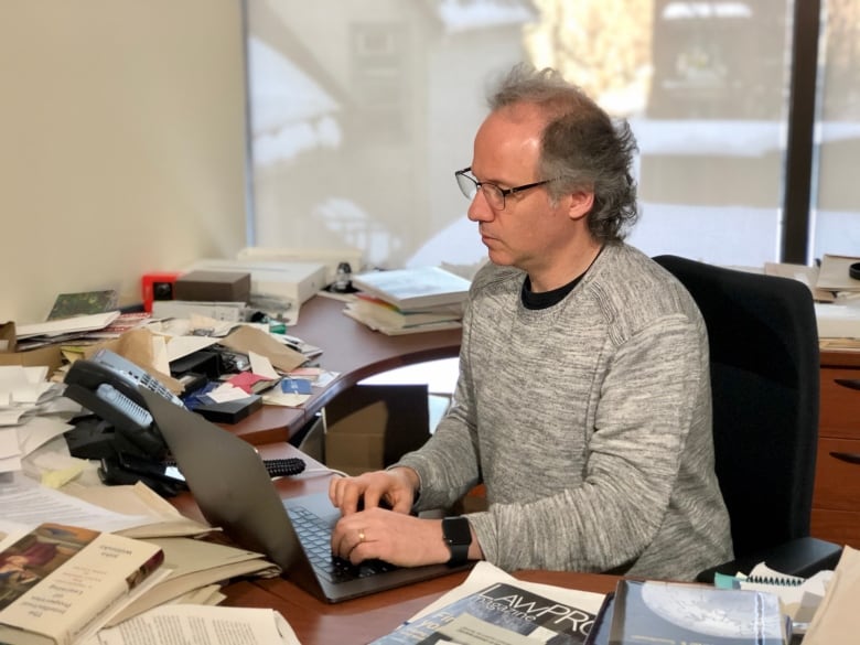 A man in a grey sweater, with papers and books covering the desk he's sitting at, looks at a laptop computer screen.