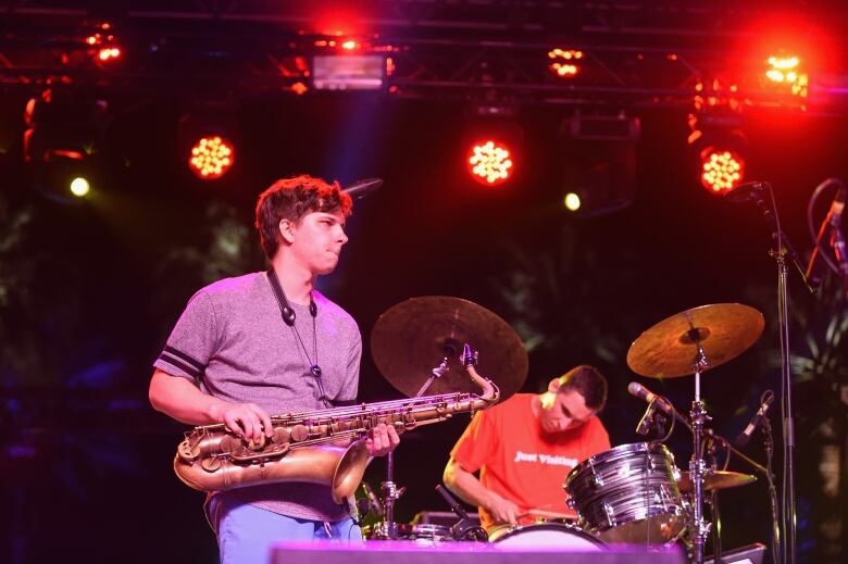 A man holding a saxophone stands in front of a man sitting at a drumset on a stage.