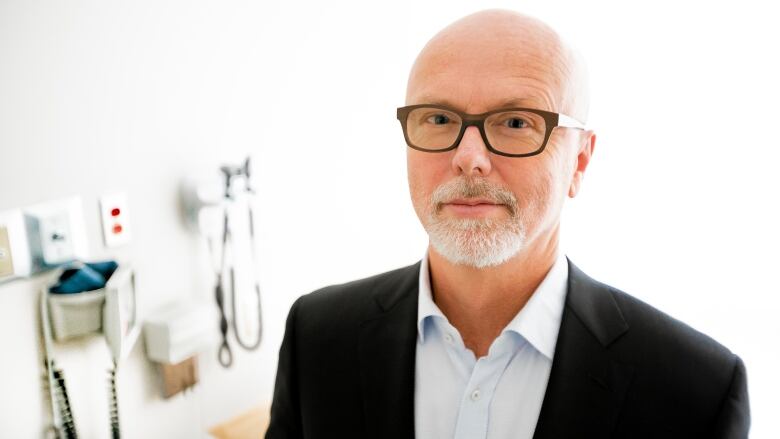 A bearded man with glasses stands in a doctors office looking at the camera.