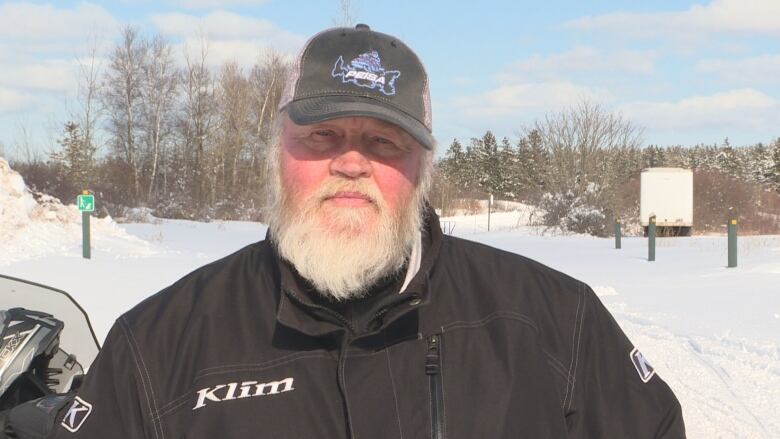 Dale Hickox stands in a ball cap and black jacket. There is snow on the ground and trees behind him. He has a white beard. 