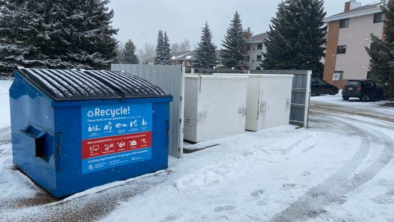 Large blue commercial recycling bin sits near snow-covered road. Spruce trees and an apartment building are in the background