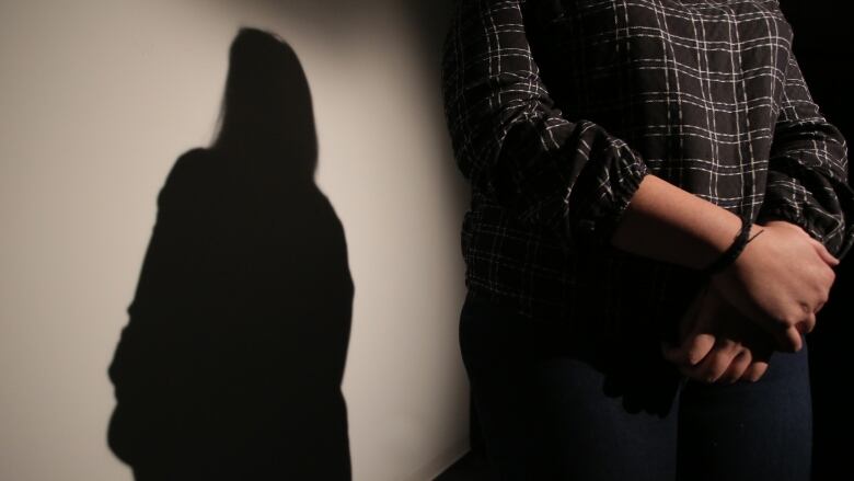 A woman stands in a darkened room. She is seen from the midsection down, her arms lowered and hands clasped.