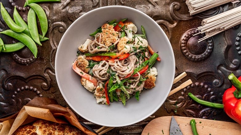 Overhead shot of a bowl of soba noodle salad with roasted cauliflower, slices of red bell pepper and sliced green beans in it. The bowl sits on a dark brown metal surface. 