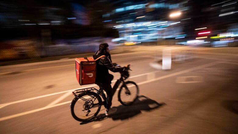 A cyclist with an orange DoorDash delivery bag bikes at night down a Vancouver street.