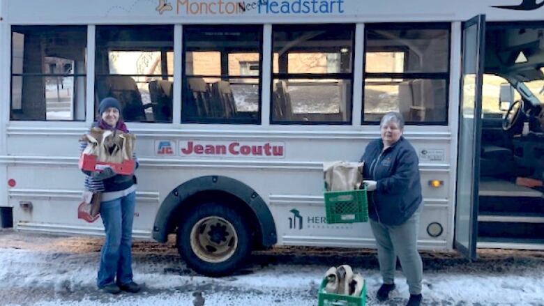 Two women stand in front of a bus handing out school lunches.