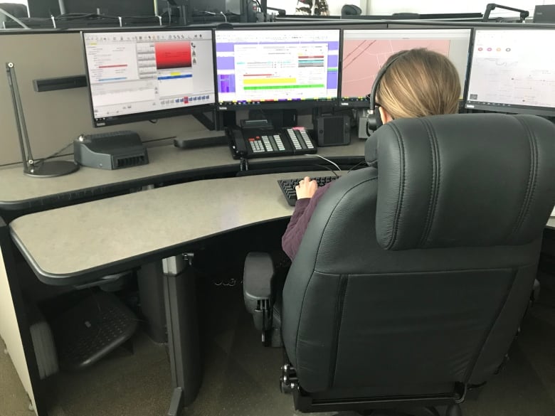 A woman is seated in a chair with headphones on, while looking at four computer monitors to help her respond to emergencies. 