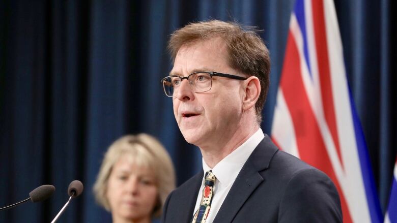 B.C. Health Minister Adrian Dix stands in front of a podium while Dr. Bonnie Henry stands in the blurry background.