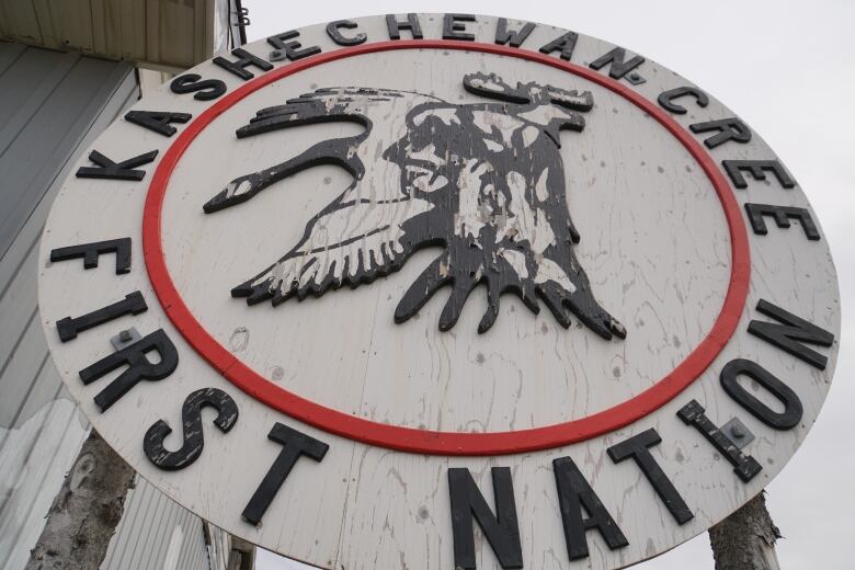 A circular white wooden sign with paint peeling off of it reads 'Kashechewan Cree First Nation' with a logo in the middle depicting a man's head, a goose and a moose.