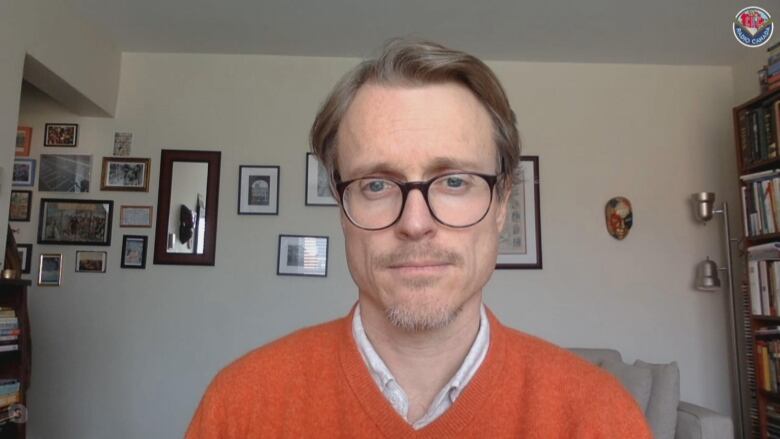  man in an orange sweater and glasses sits ina living room with pictures on the wall behind him