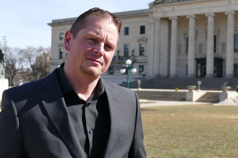 A man wearing a black suit stands in front of the Manitoba Legislative building.