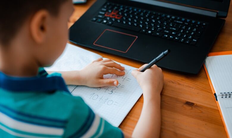 Looking over shoulder of young child doing math homework on a piece of paper. A laptop's keyboard and another notebook are visible