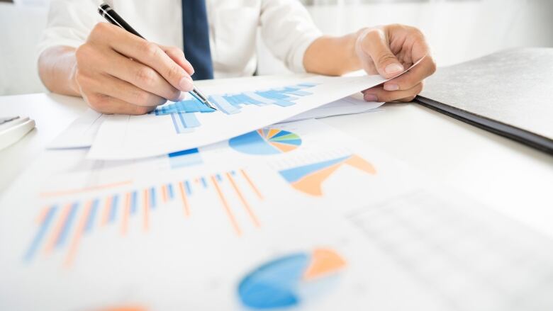 A stock photo shows a man from the shoulders down holding a pen and leaning over a stack of white papers with colourful pie graphs