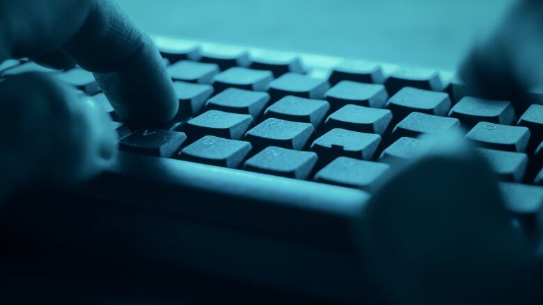 A closeup shows the hands of a person typing on a keyboard in a darkened room.