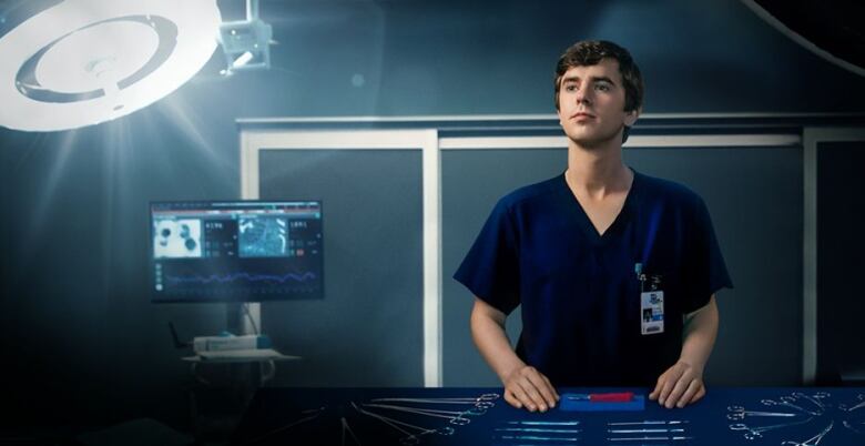 A promotional shot for the medical drama The Good Doctor shows a young, white man in scrubs standing next to a bed in a hospital room with medical instruments laid out in front of him on a steel tray. 