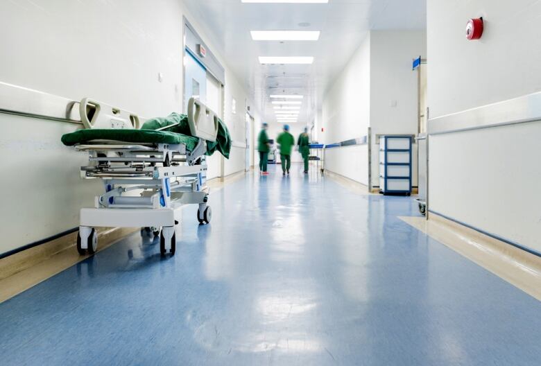 An empty gurney in the hallway of a hospital. Doctors in green scrubs stand in the background. 