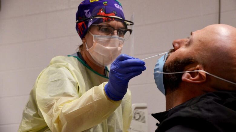 A bald man with a beard leans his head back as a woman wearing a mask and protective gown shoves a swab up his nose