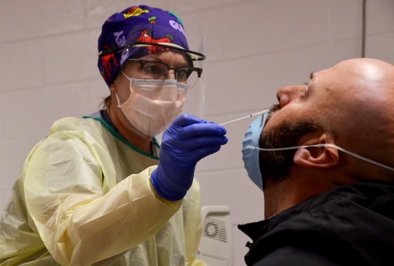 A bald man with a beard leans his head back as a woman wearing a mask and protective gown shoves a swab up his nose