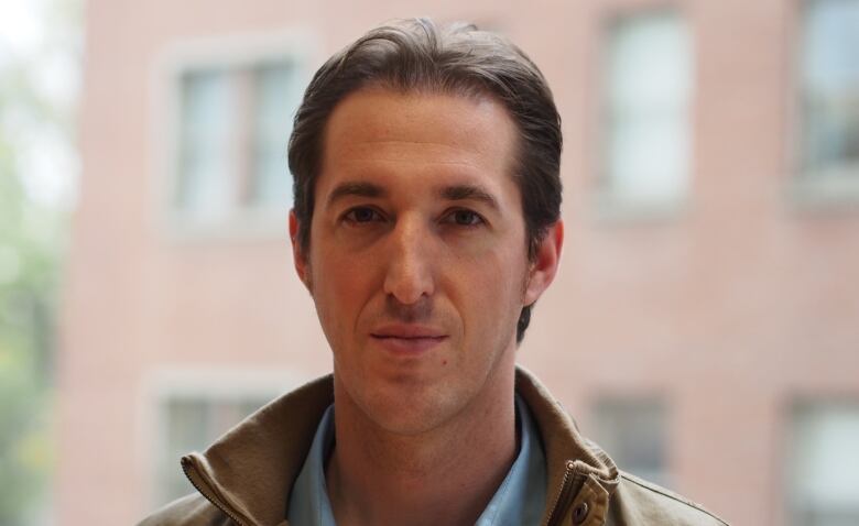 A man with short brown hair slightly smiles for a photo. He's wearing a jacket and a blue button-up shirt.