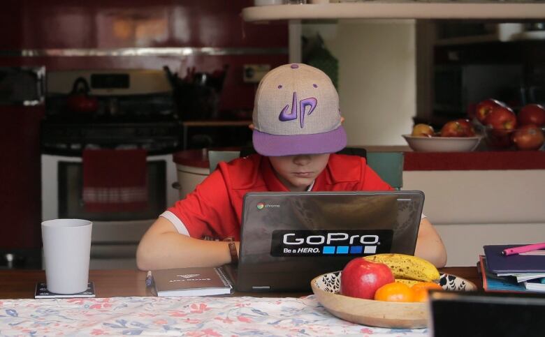 A kid in a red t-shirt and ballcap pulled low over the eyes works at a laptop at a kitchen table. 