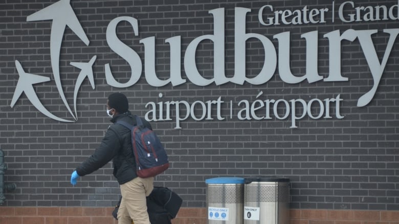 A man wearing a mask walking in front of a building that says Greater Sudbury Airport.