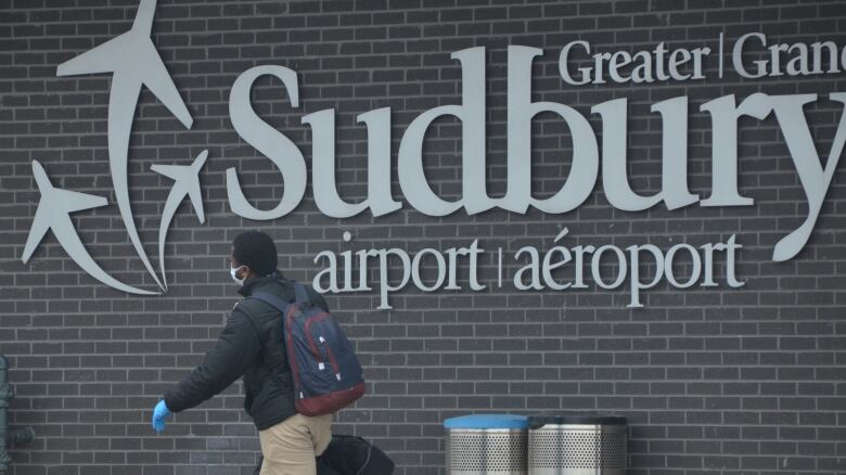 A man wearing a mask walking in front of a building that says Greater Sudbury Airport.