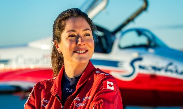 A smiling woman in a red jumpsuit stands in front of a Snowbird training plane.