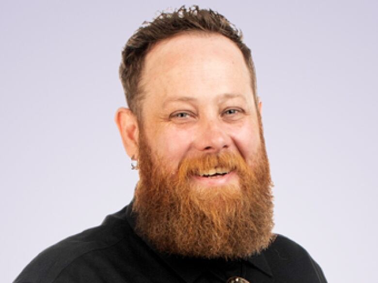 Caucasian man with brown hair and a long red beard smiles at the camera. SHot is from the shoulders up, he's wearing a black dress shirt