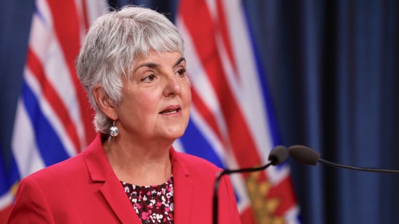 A woman with short white hair, wearing a red blazer speaks at a rostrum.